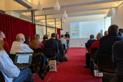 Ein Mann an einem Podium hält einen Vortrag vor Zuhörenden.