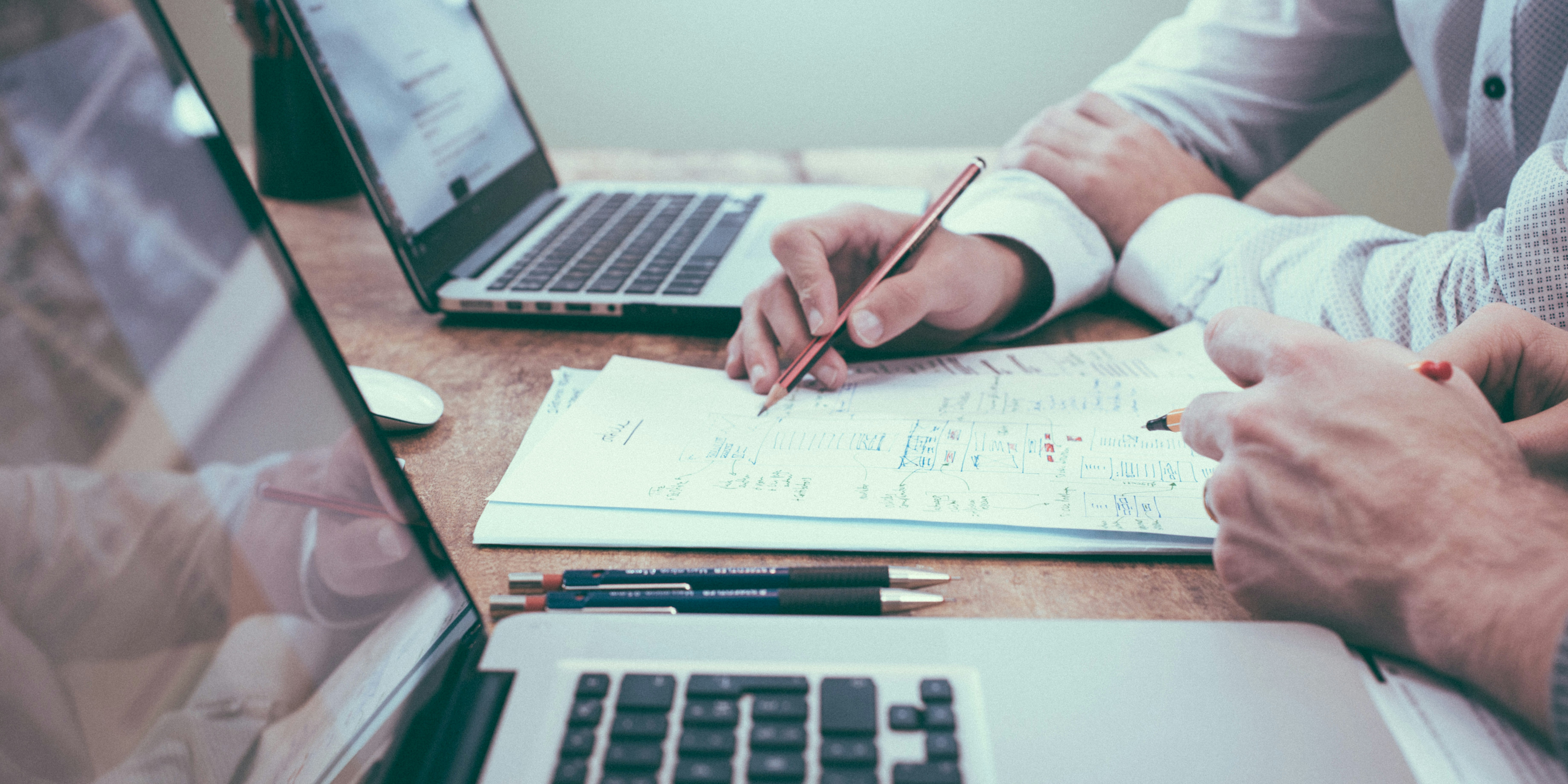 Zwei Laptops stehen aufgefklappt auf einem Arbeitstisch. Dazwischen liegen ausgedruckte Diagramme an denen zwei Personen handschriftliche Notizen machen. Von den Personen sind nur die Hände und Teile der Arme im Bild.