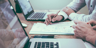 Zwei Laptops stehen aufgefklappt auf einem Arbeitstisch. Dazwischen liegen ausgedruckte Diagramme an denen zwei Personen handschriftliche Notizen machen. Von den Personen sind nur die Hände und Teile der Arme im Bild.