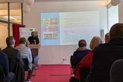 Ein Mann an einem Podium hält einen Vortrag vor Zuhörenden.