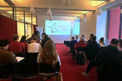 Ein Vortragsraum von hinten aufgenommen. Mehrere besetzte Sitzreihen und vorne ein Sprecher vor einer Präsentation "Willkommen! Kioskwelten 2025".