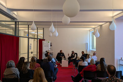 Ein Vortragsraum von hinten aufgenommen mit mehreren besetzten Stuhlreihen. Vorne sitzen eine Frau und drei Männer auf großen Sesseln und diskutieren.