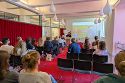 Ein Vortragsraum von hinten aufgenommen mit mehreren besetzten Stuhlreihen. Am Pult stehen zwei Studierende und halten einen Vortrag.