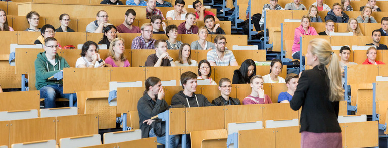 Eine Dozierende steht vor Studierenden in der Vorlesung im Hörsaal. 