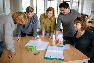 Fünf Studierende im Seminarraum stehen um einen Tisch herum und sehen auf ein Dokument